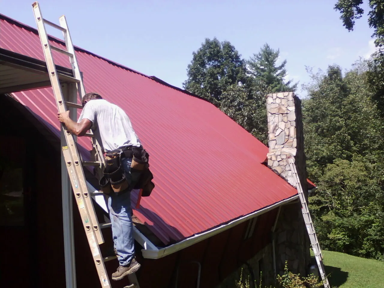 Roofer on a ladder beside a red metal roof and stone chimney — Soffit & Fascia Repair by Roofing Brothers in Veyo